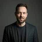 Portrait of a man with beard wearing a dark shirt looking at camera.